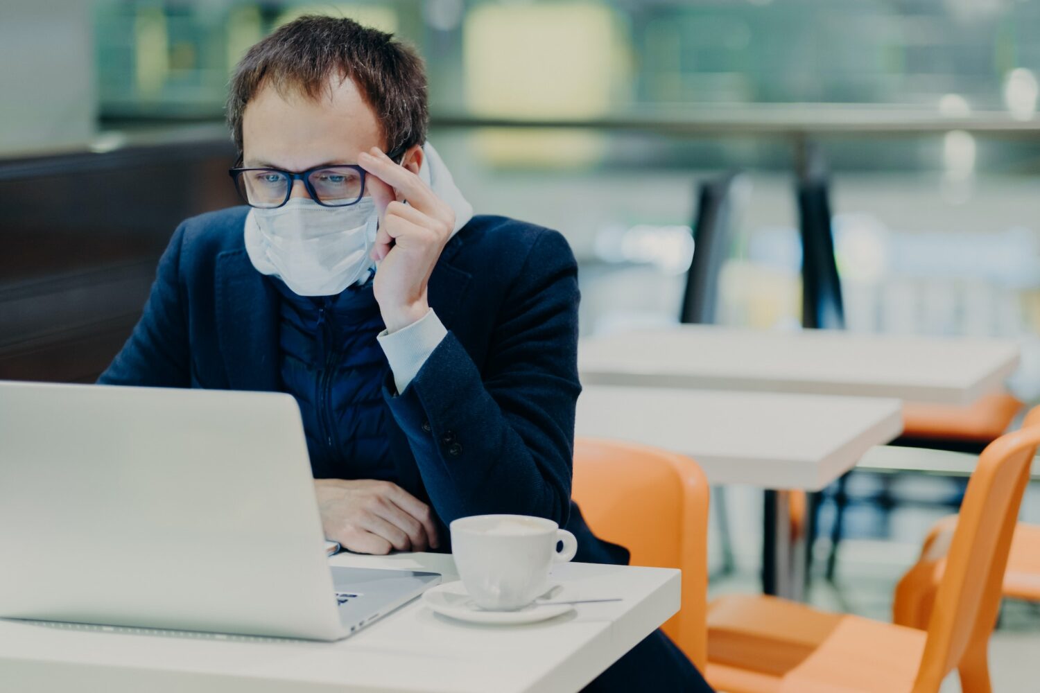 Man in medical mask and spectacles, sits in front of modern laptop computer, poses in cozy cafeteria